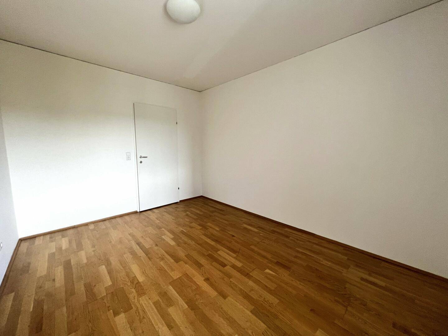 Helles, leeres Zimmer mit Parkettboden und weißer Tür, ideal als Schlaf- oder Arbeitszimmer.