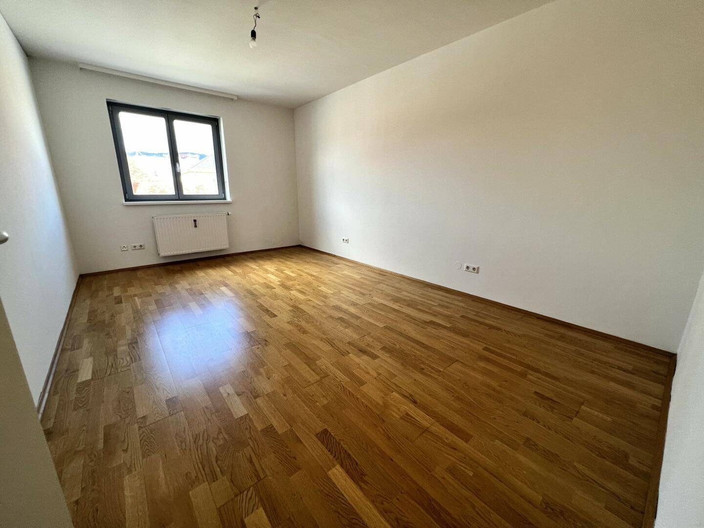 Geräumiges, leeres Zimmer mit Parkettboden und Fenster, ideal für Schlaf- oder Arbeitsbereich.