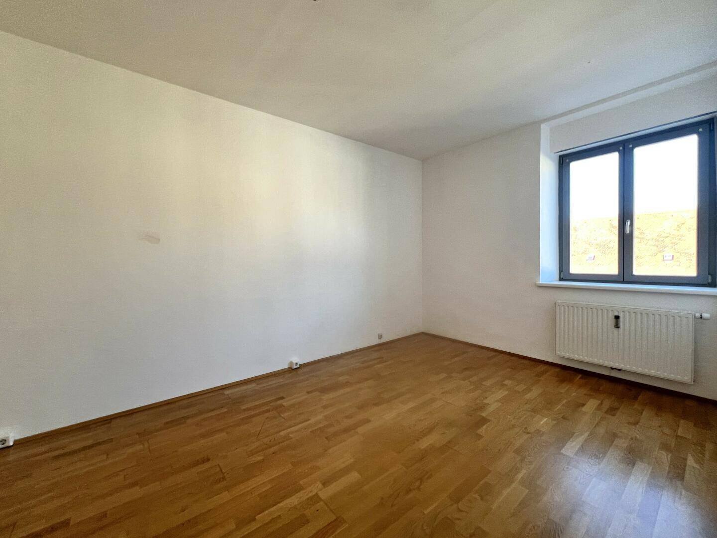 Heller, leerer Raum mit Parkettboden und großem Fenster, ideal als Wohn- oder Schlafzimmer.