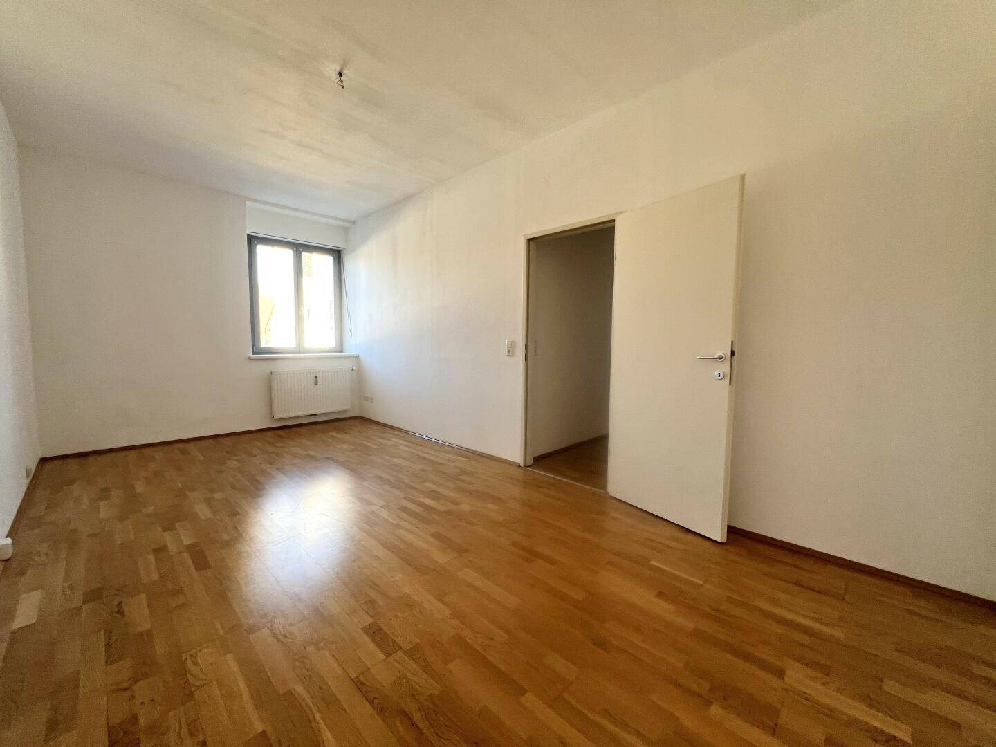 Geräumiger, leerer Raum mit Parkettboden und Zugang zu einem weiteren Zimmer, in gutem Zustand.