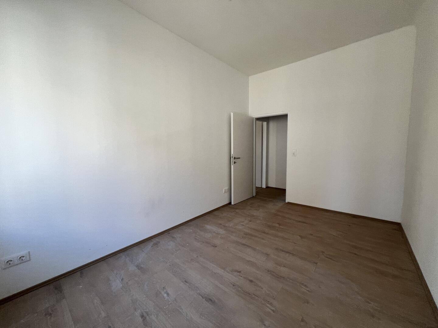 Helles Zimmer mit Laminatboden und weiß gestrichenen Wänden, mit Zugang zu einem weiteren Raum.