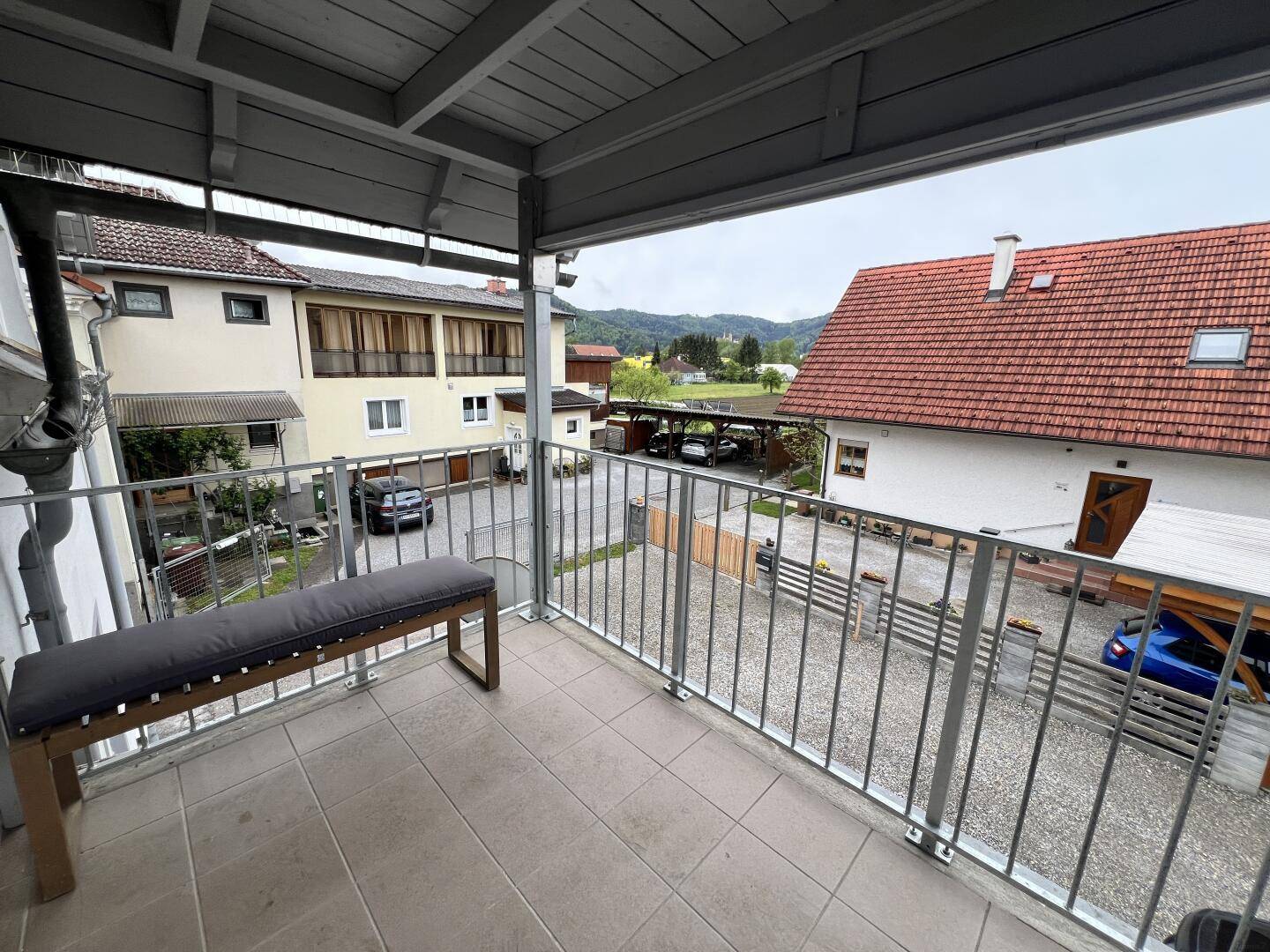 Überdachter Balkon mit Sitzbank und Blick auf die Nachbarschaft sowie grüne Hügel.