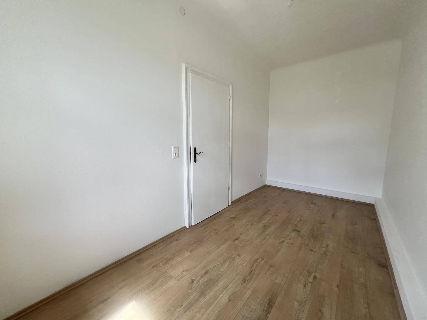 Leeres Zimmer mit Laminatboden und weiß gestrichenen Wänden, bereit zur individuellen Gestaltung.