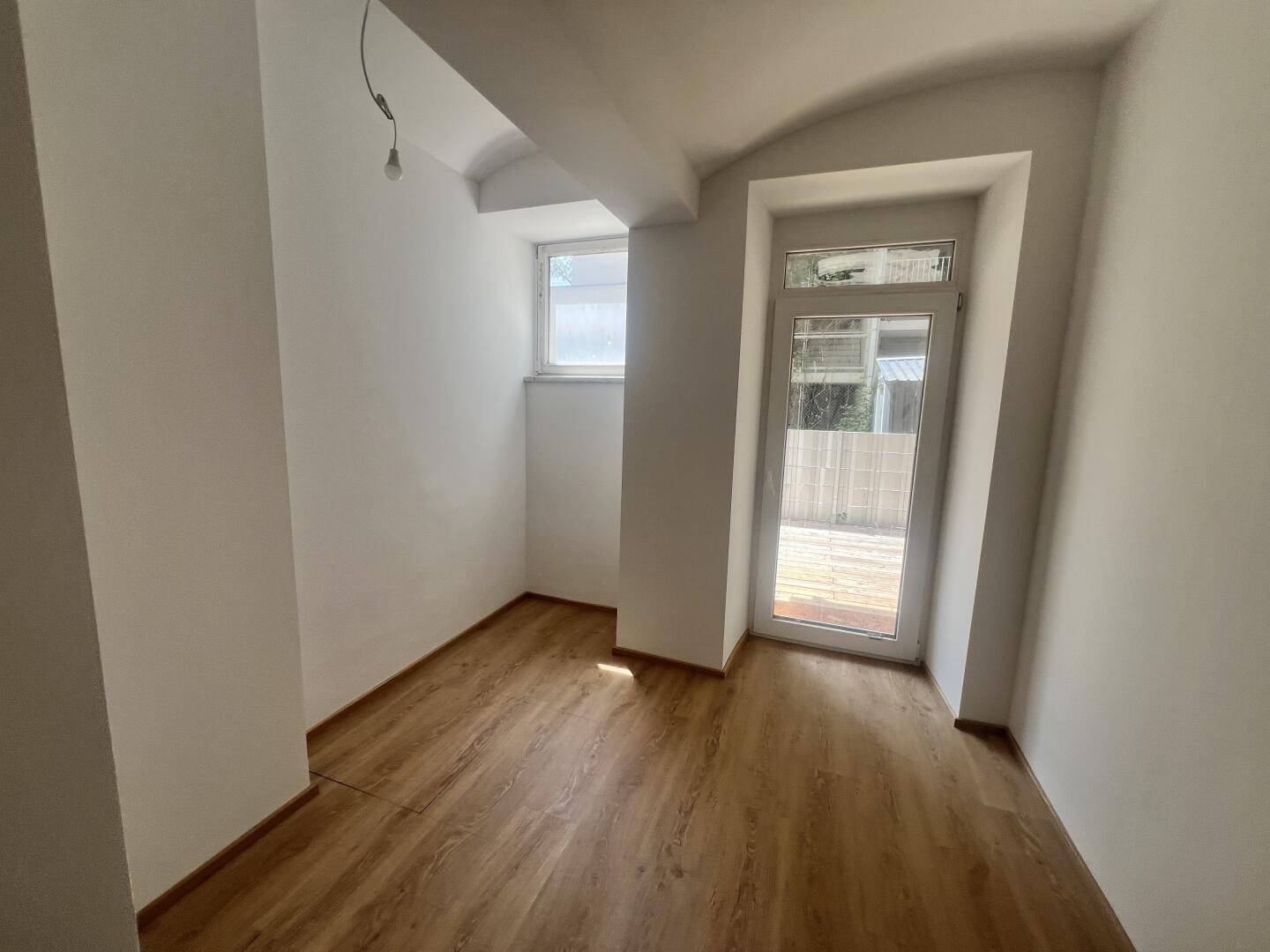 Helles Zimmer mit Laminatboden, Fenster und Zugang zu einem kleinen Außenbereich.