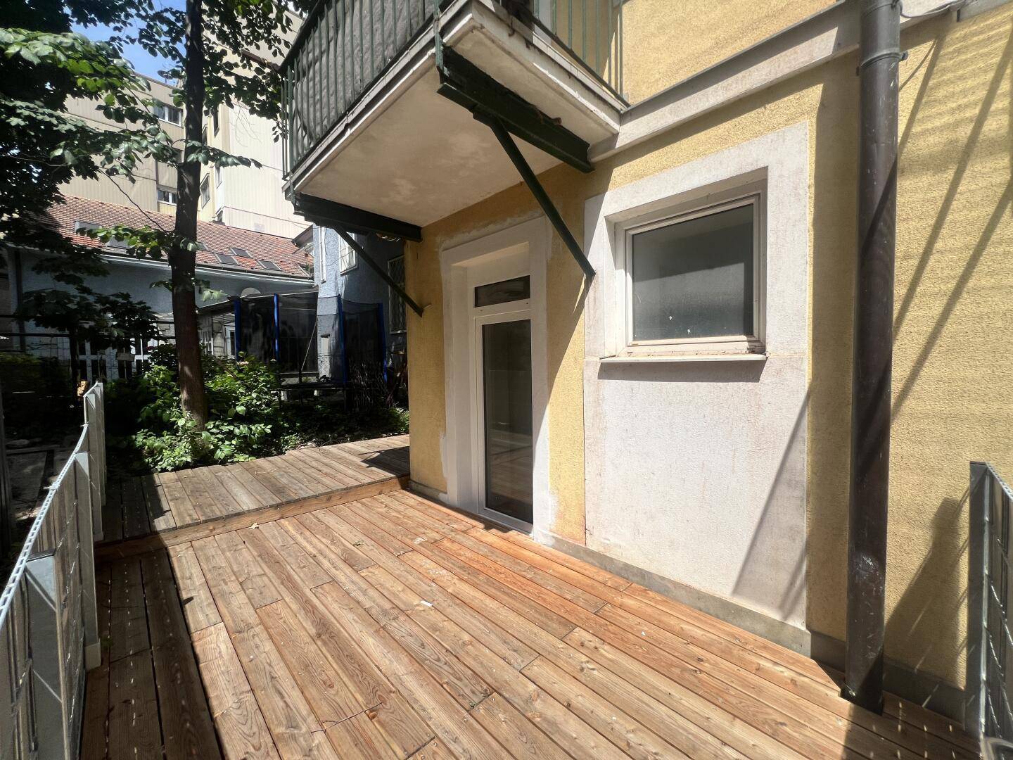 Sonnige Holzterrasse mit Blick auf die Gebäudefassade und angrenzende Grünflächen.