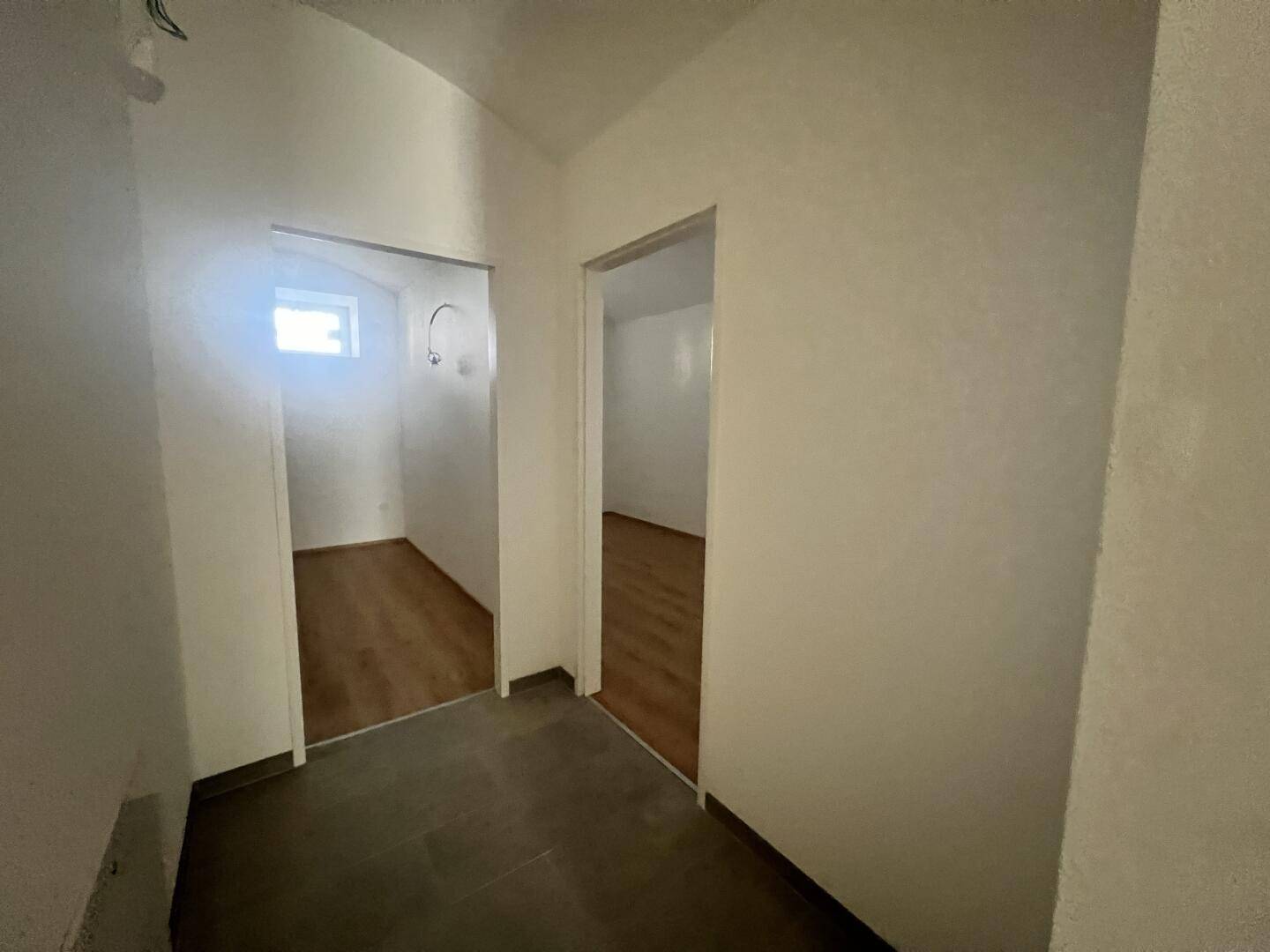 Schmaler Flur mit grauen Fliesen, der zu zwei Zimmern mit Holzboden und einem kleinen Fenster führt.