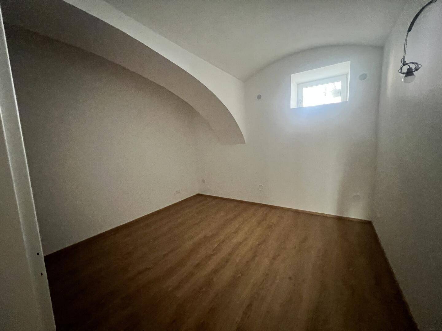 Leeres Zimmer mit Laminatboden, weißen Wänden, einem Gewölbedecke und einem kleinen Fenster.