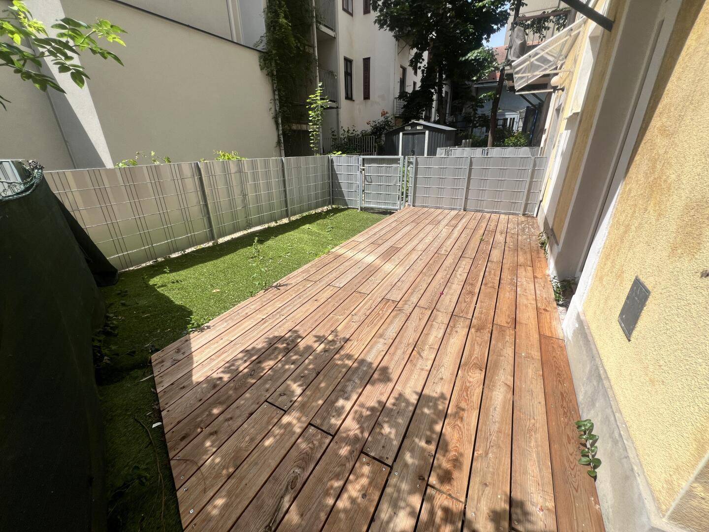 Sonnenbeschienene Holzterrasse mit angrenzendem Rasenbereich und Zaun.