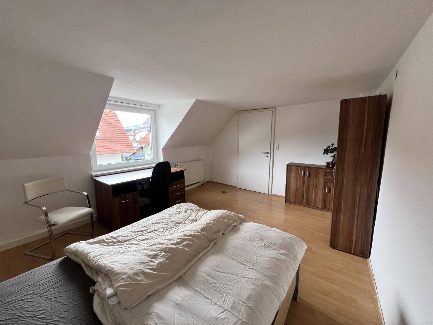 Geräumiges Schlafzimmer mit Dachschrägen, Holzboden und Blick auf die umliegenden Häuser und Dächer.