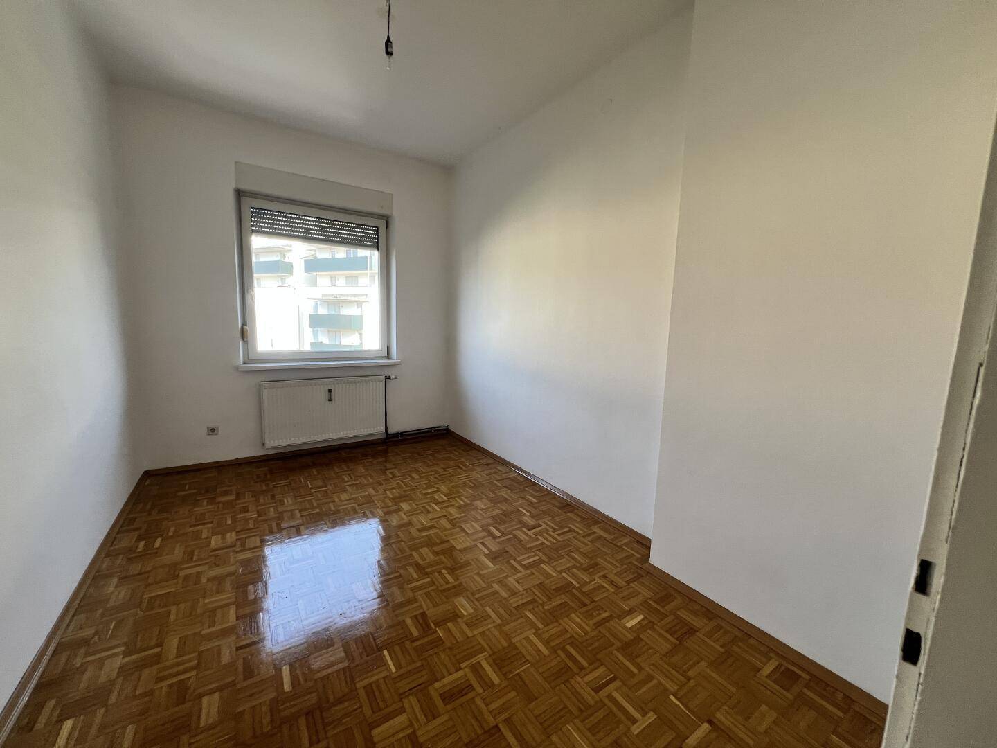 Helles Zimmer mit Parkettboden und Fenster mit Rollladen, ideal als Schlaf- oder Arbeitszimmer.