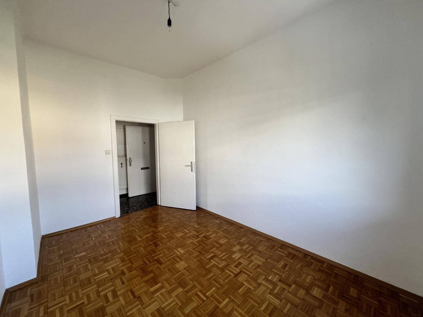 Geräumiges Zimmer mit Parkettboden und Zugang zu einem weiteren Bereich, möglicherweise Flur oder Küche.