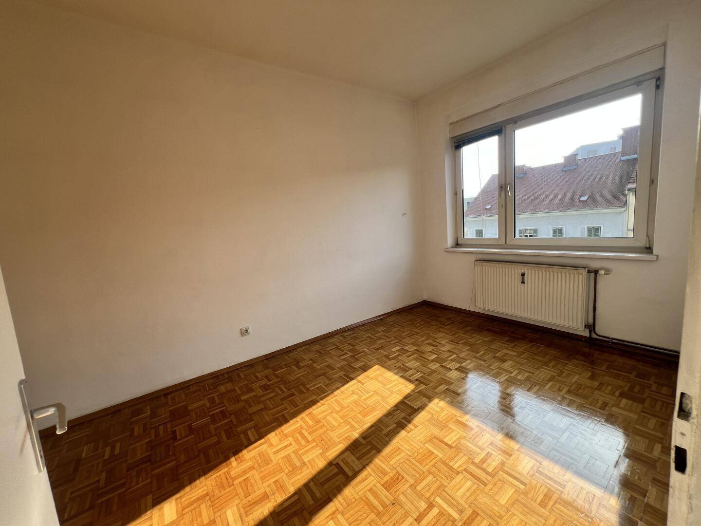 Sonniges Zimmer mit Parkettboden, weißen Wänden und einem großen Fenster.