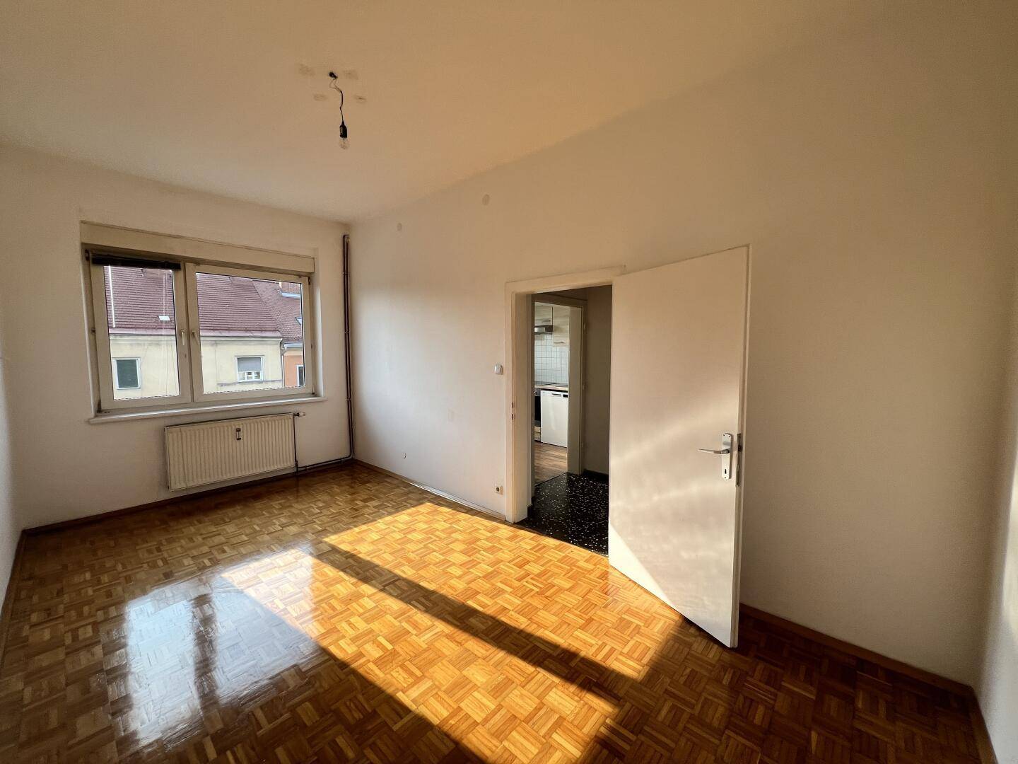 Geräumiges Zimmer mit Parkettboden und Fenster, das zu einem Flur führt.