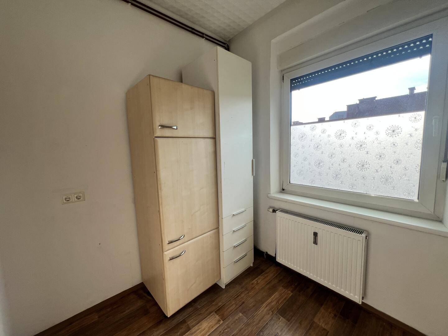 Küchenbereich mit einem hohen Schrank, Fenster und Laminatboden.