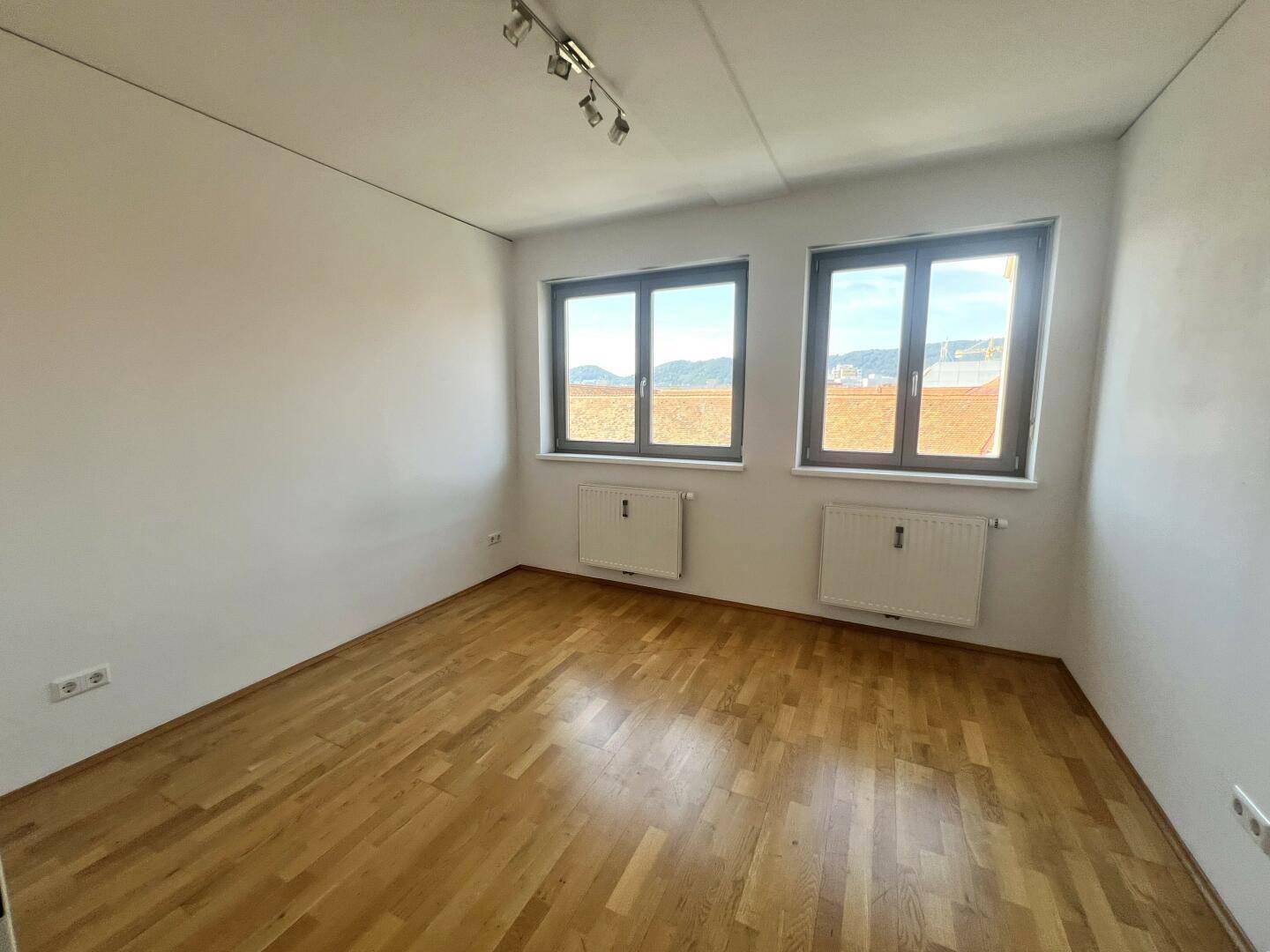 Leeres Zimmer mit Holzboden, weißen Wänden und zwei Fenstern.