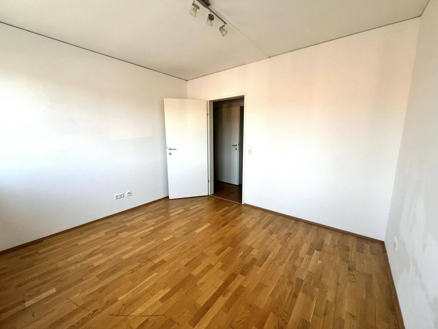 Leeres Zimmer mit Holzboden, weißen Wänden und offener Tür.