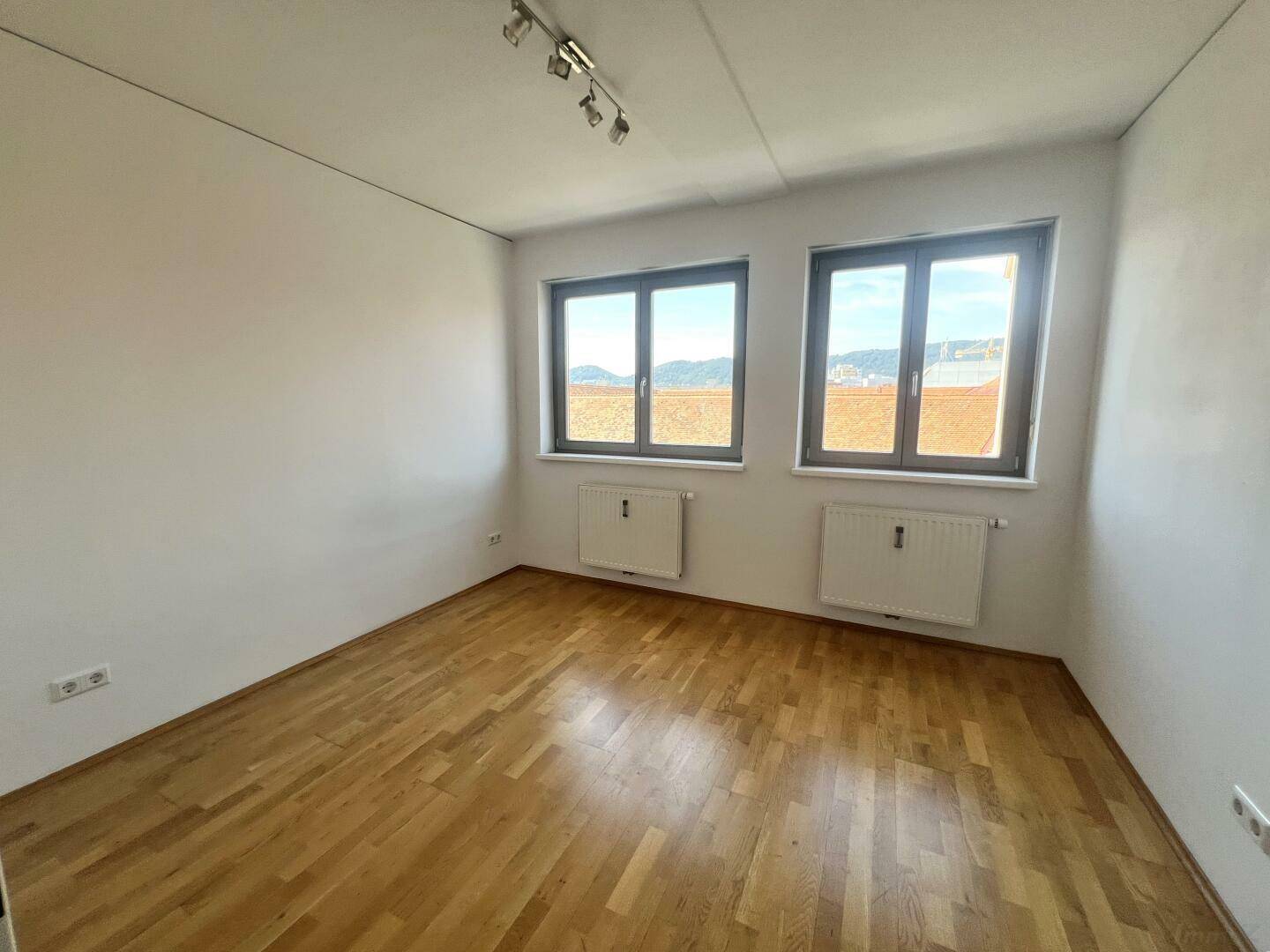 Leeres Schlafzimmer mit hellem Parkettboden und zwei Fenstern, die viel Tageslicht hereinlassen.