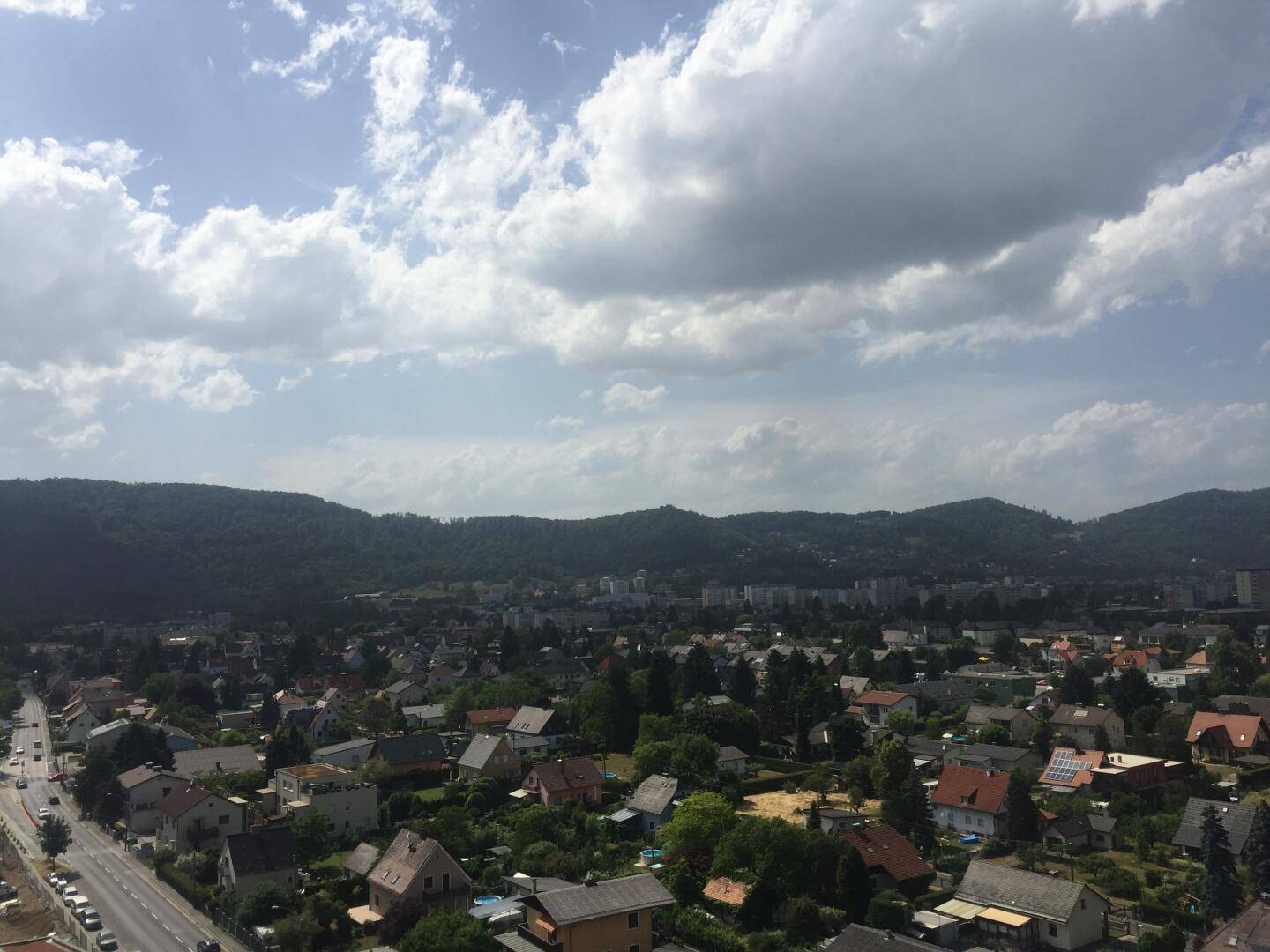 Weitläufiger Blick über die Stadt mit Wohngebäuden und bewaldeten Hügeln im Hintergrund.