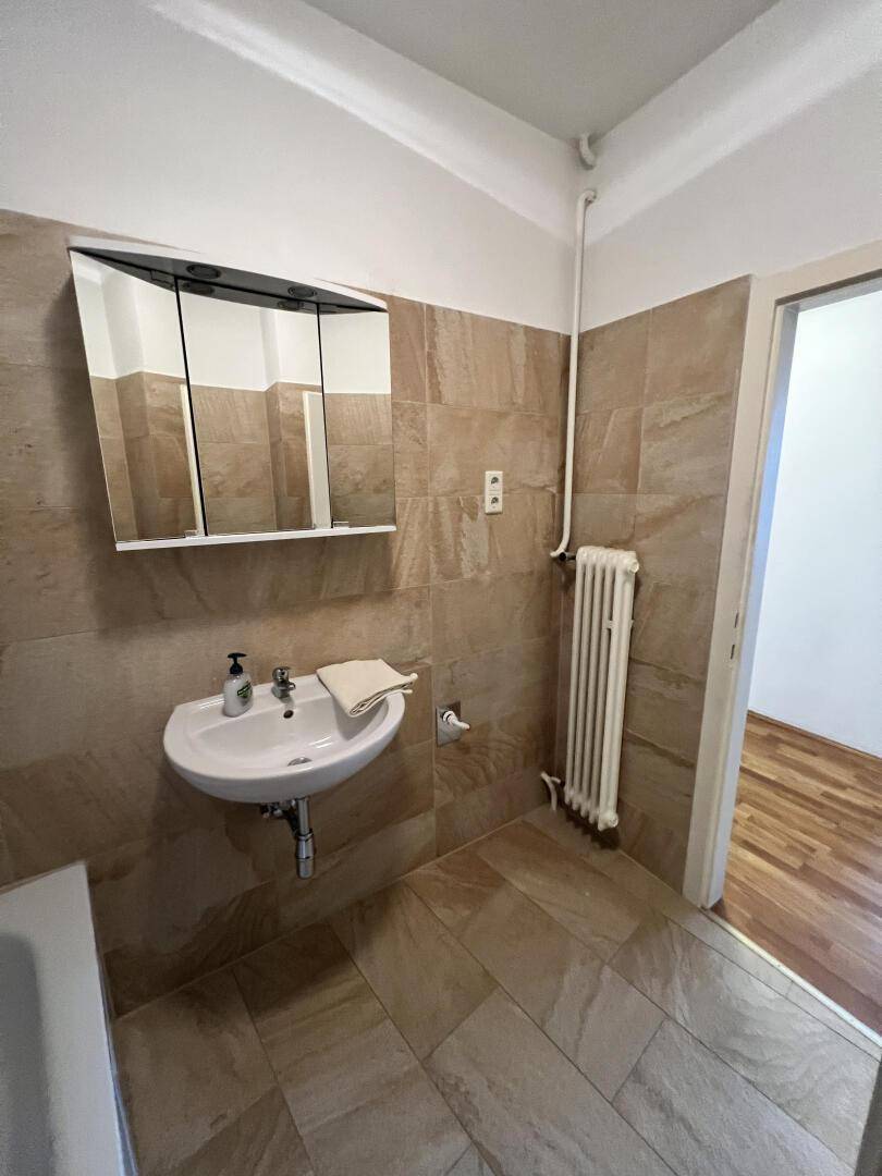 Badezimmer mit Waschbecken, Spiegelschrank und Heizkörper, gefliest in beigen Farbtönen.