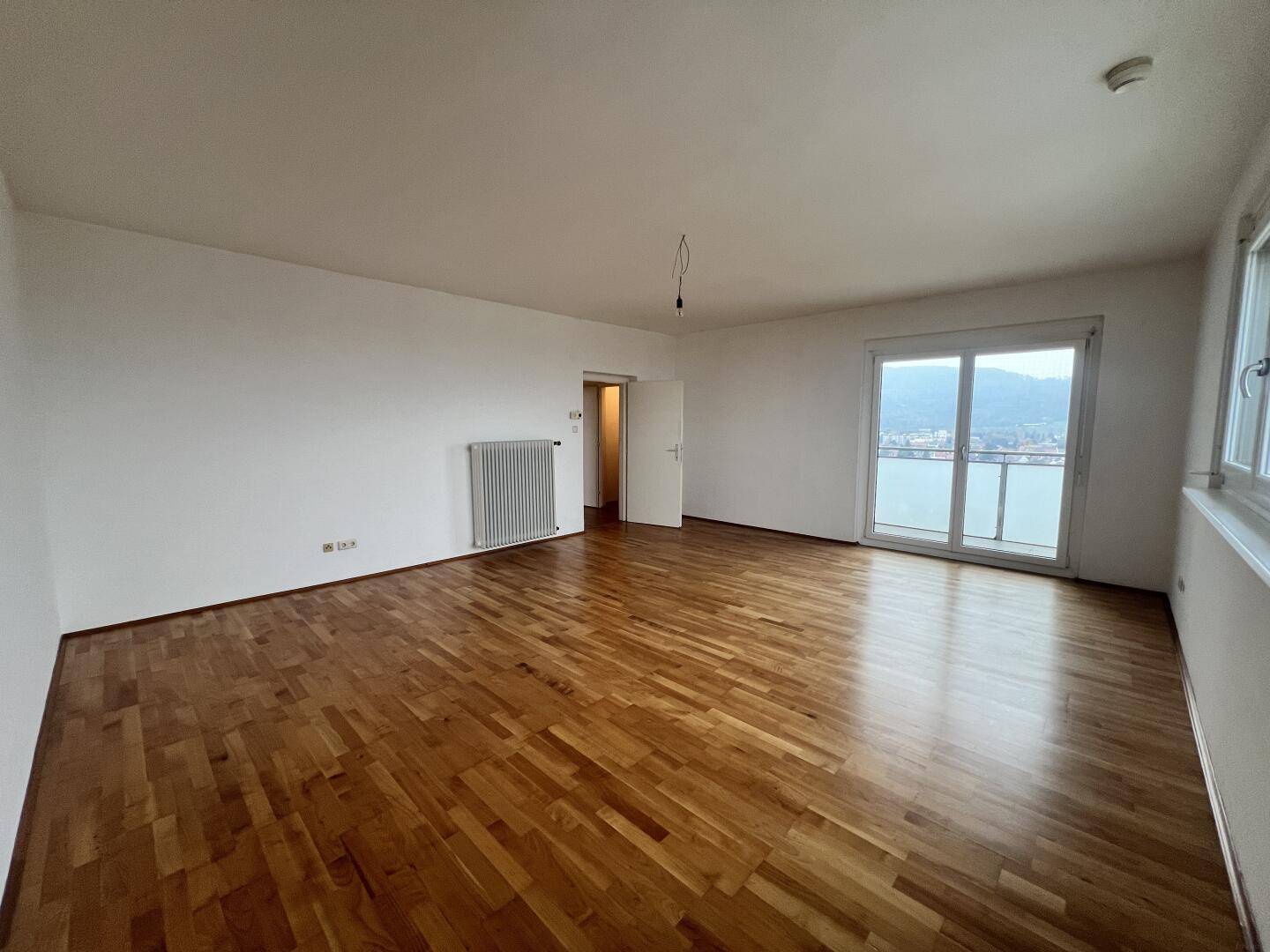 Geräumiges Wohnzimmer mit Parkettboden, Heizkörper und Zugang zu einem weiteren Raum.