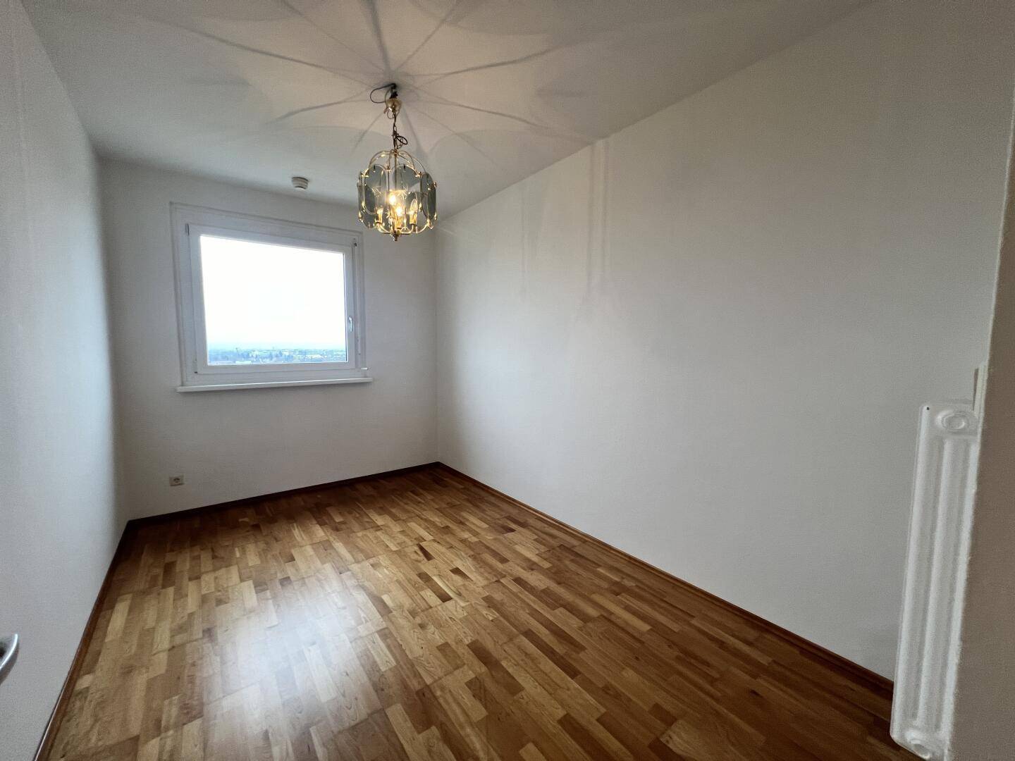 Helles Zimmer mit Parkettboden, Fenster und einer dekorativen Deckenleuchte.
