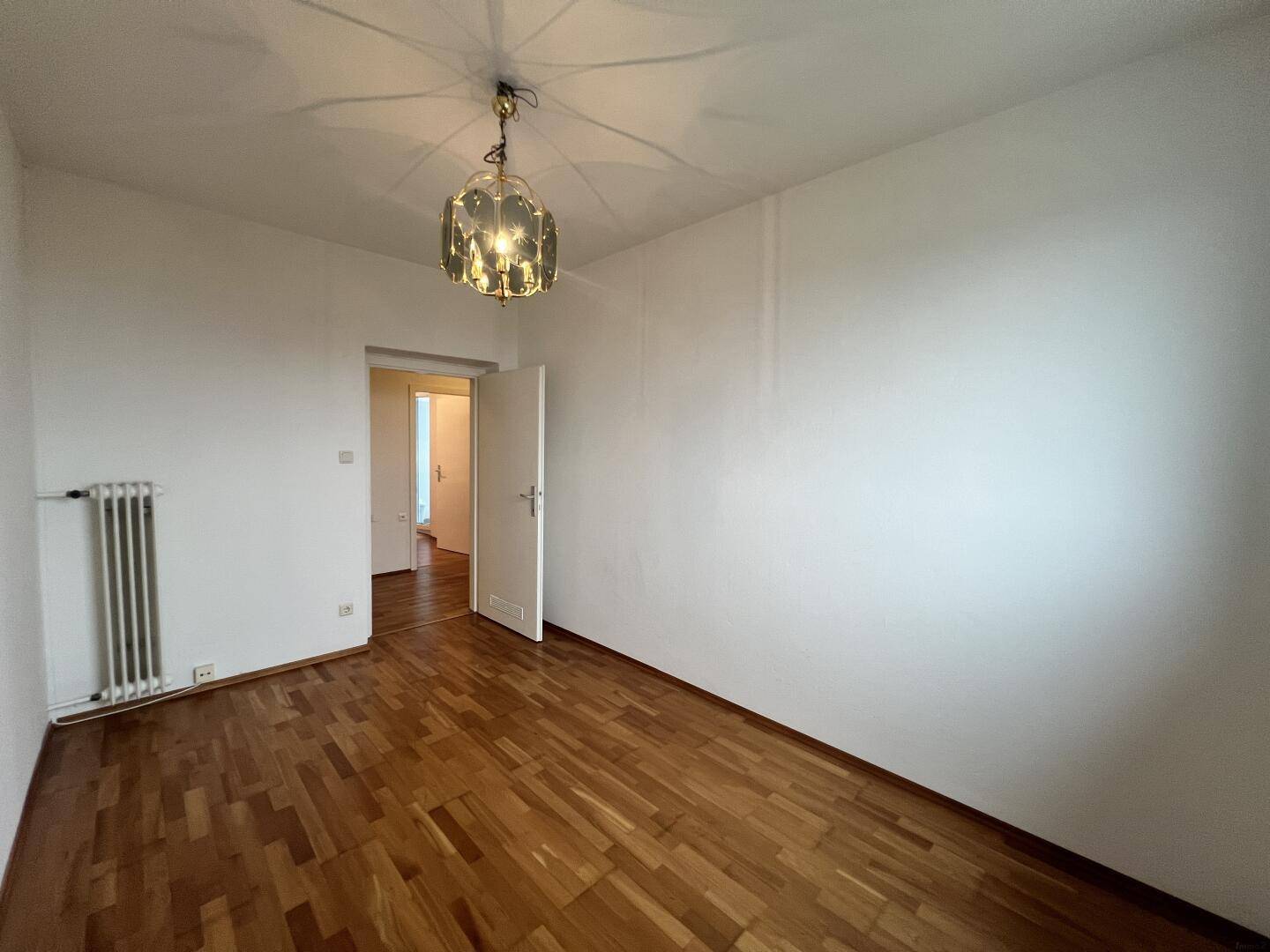 Leeres Zimmer mit Parkettboden, Heizkörper und Zugang zu einem Flur.