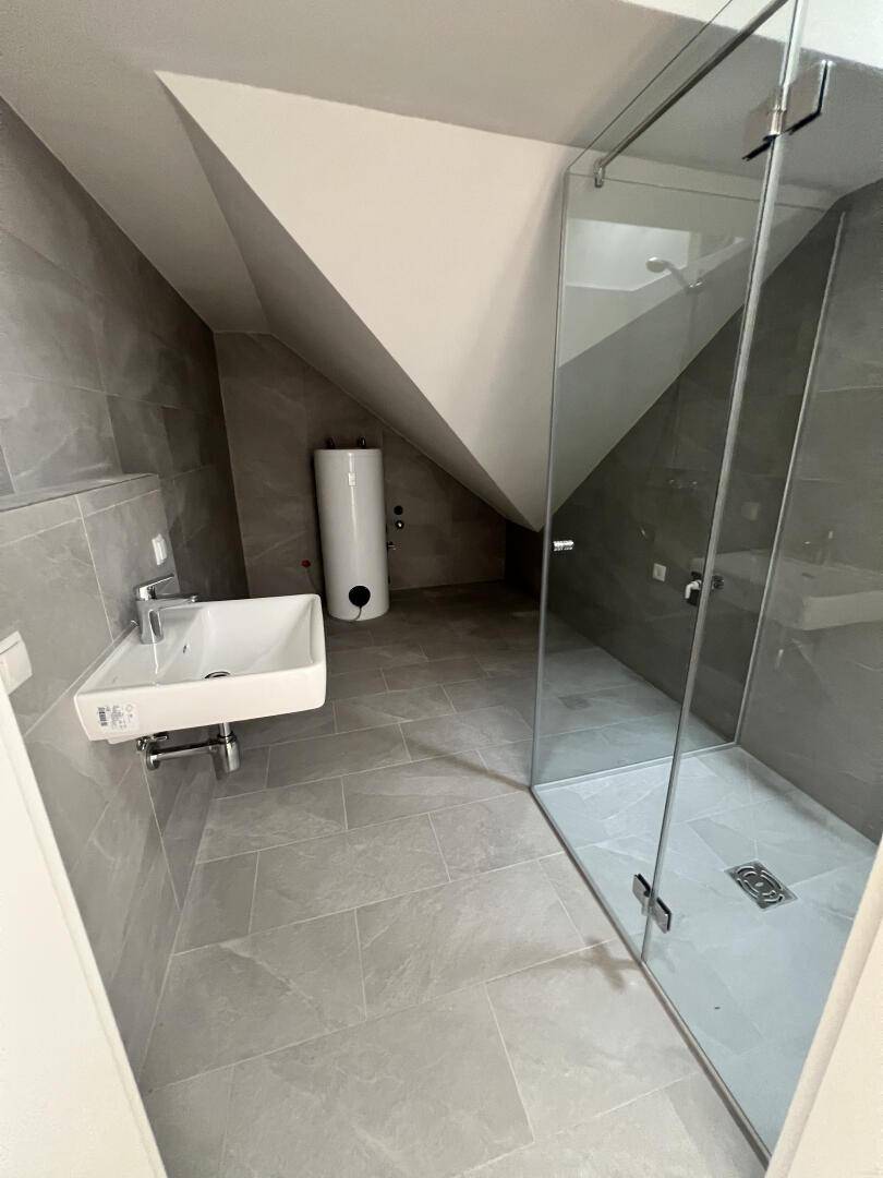 Modernes Badezimmer mit grauen Fliesen, Waschbecken und Glasdusche unter der Dachschräge.