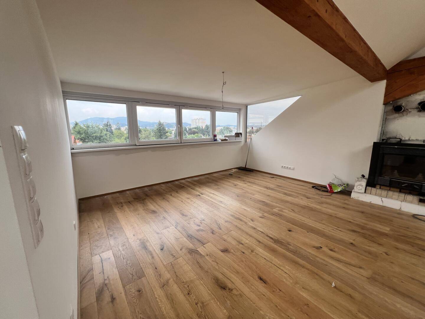 Helles Zimmer mit Holzboden, großem Fenster und vorbereitetem Kaminbereich.