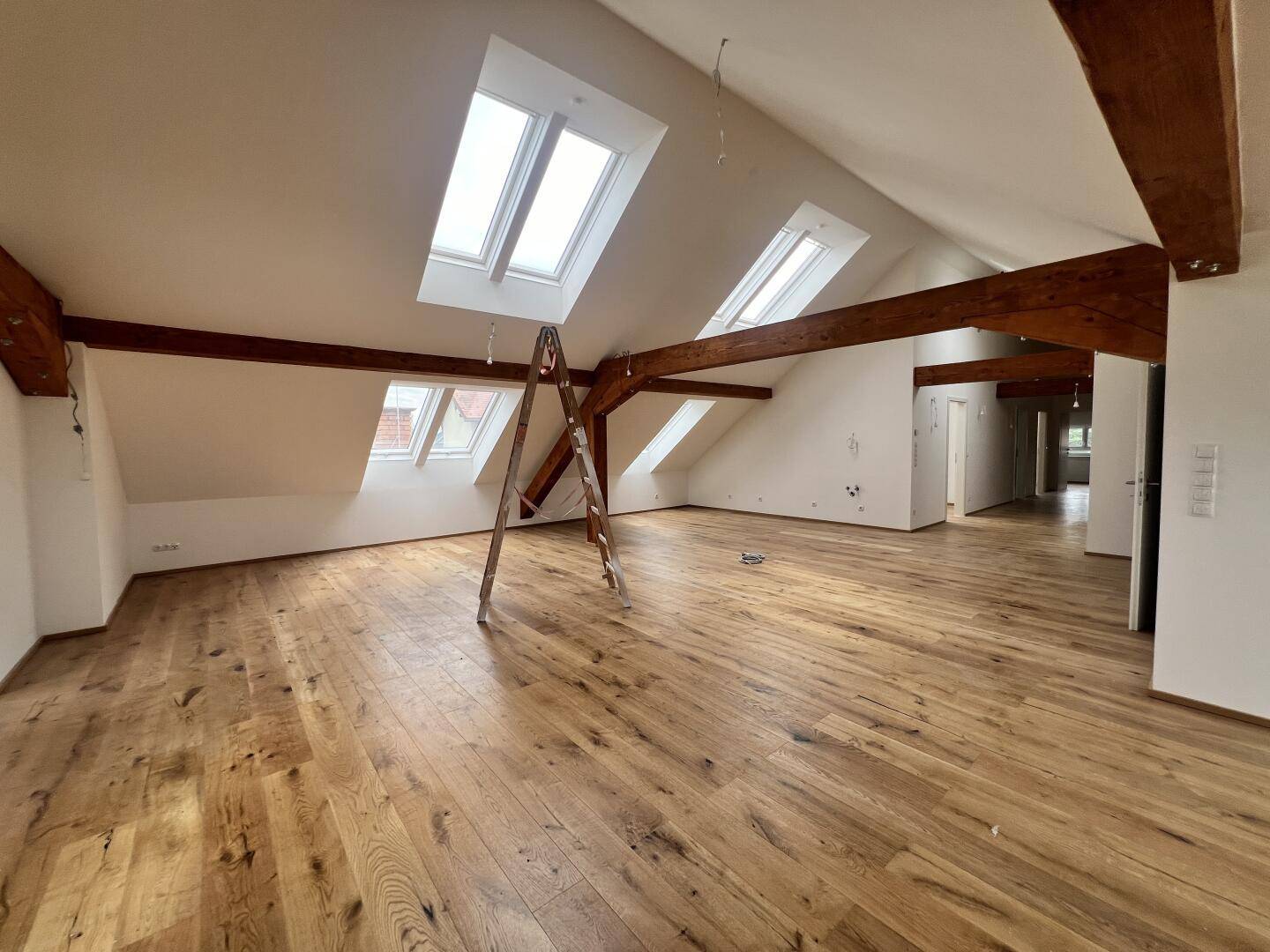 Helles Wohnzimmer mit Holzboden, sichtbaren Balken und mehreren Dachfenstern.