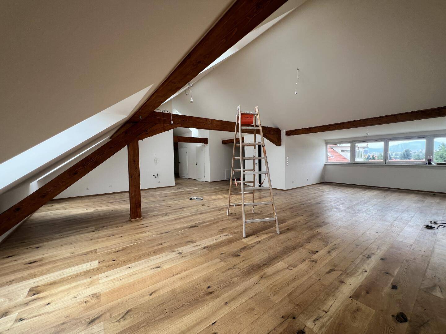 Großes Dachgeschosszimmer mit Holzboden, freiliegenden Holzbalken und Dachfenstern.