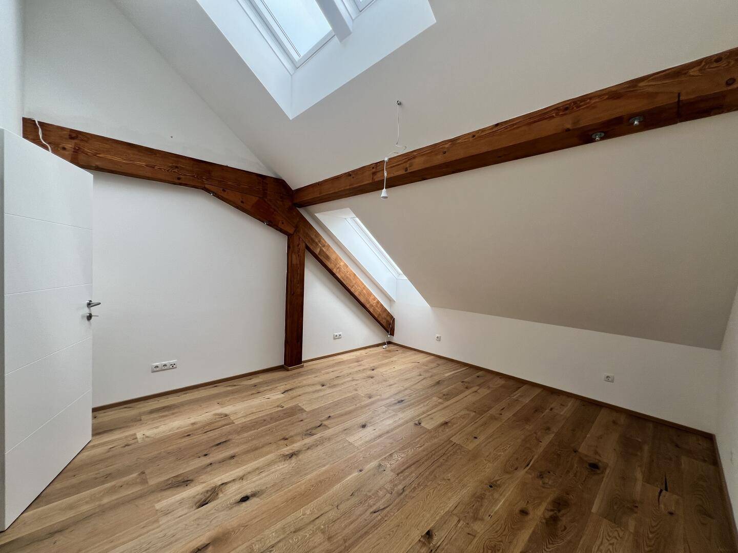 Helles Zimmer mit Holzboden, sichtbaren Balken, Dachschräge und Dachfenstern.
