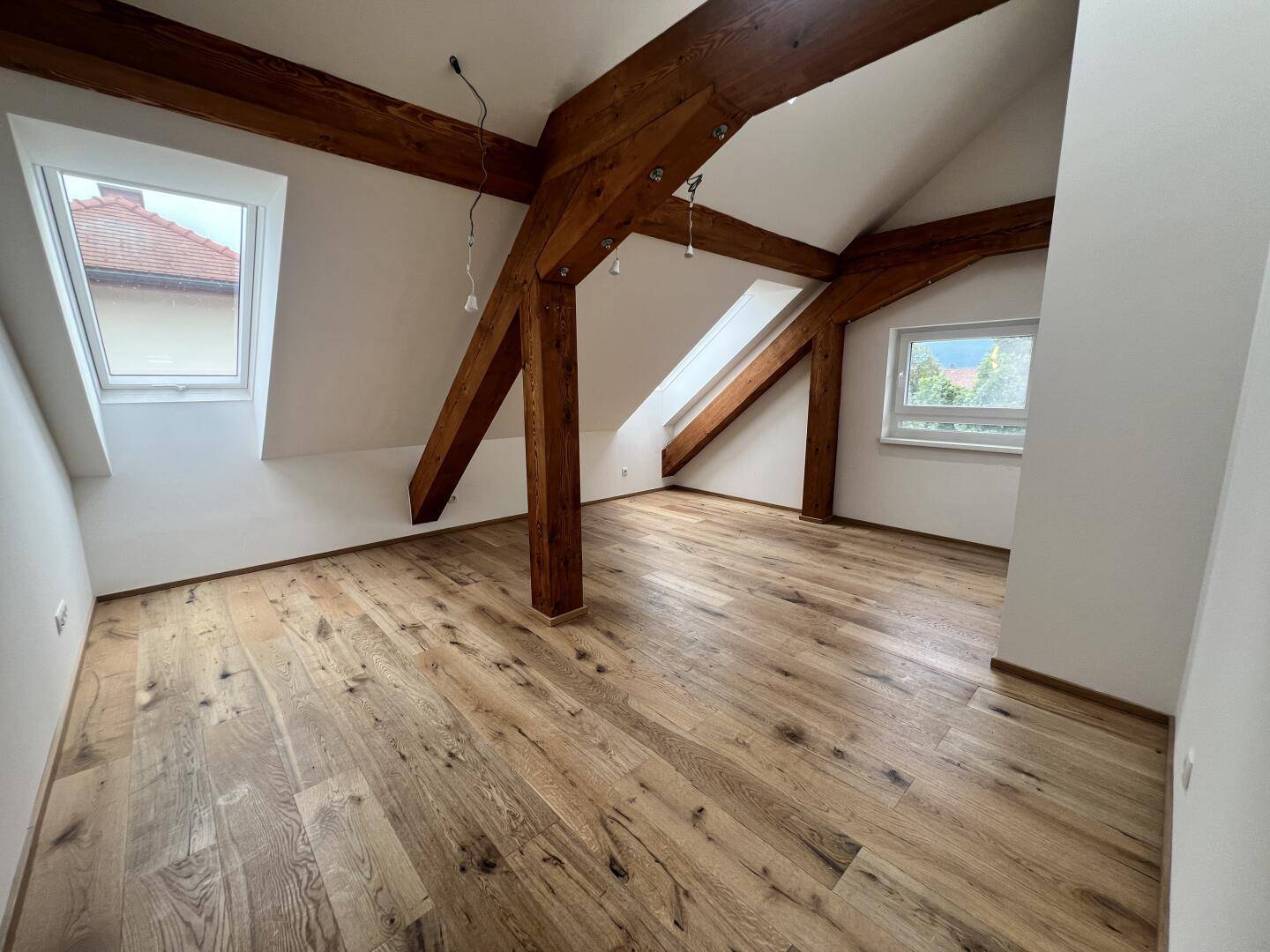 Helles Zimmer mit Holzboden, sichtbaren Balken, Dachschräge und Fenstern.