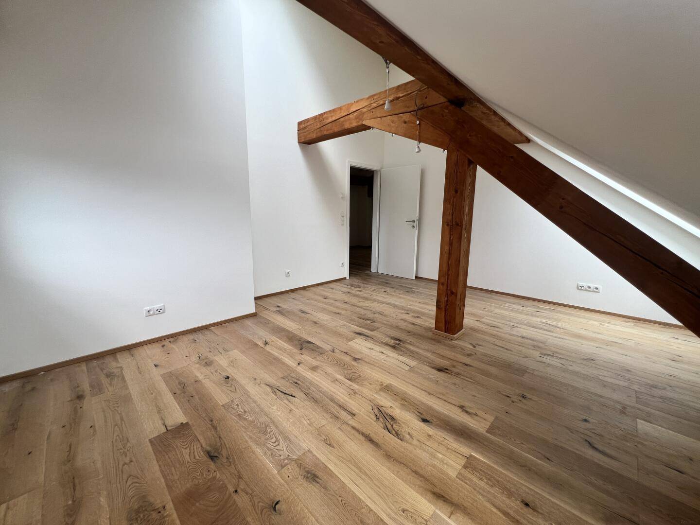 Zimmer mit Holzboden, Holzbalken, Dachschräge und weißer Tür.