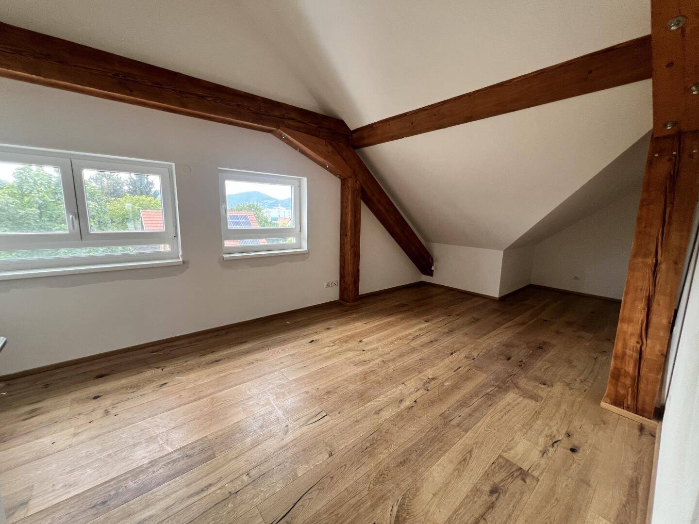 Zimmer mit Holzboden, Holzbalken, Dachschräge und Fenstern mit Ausblick.
