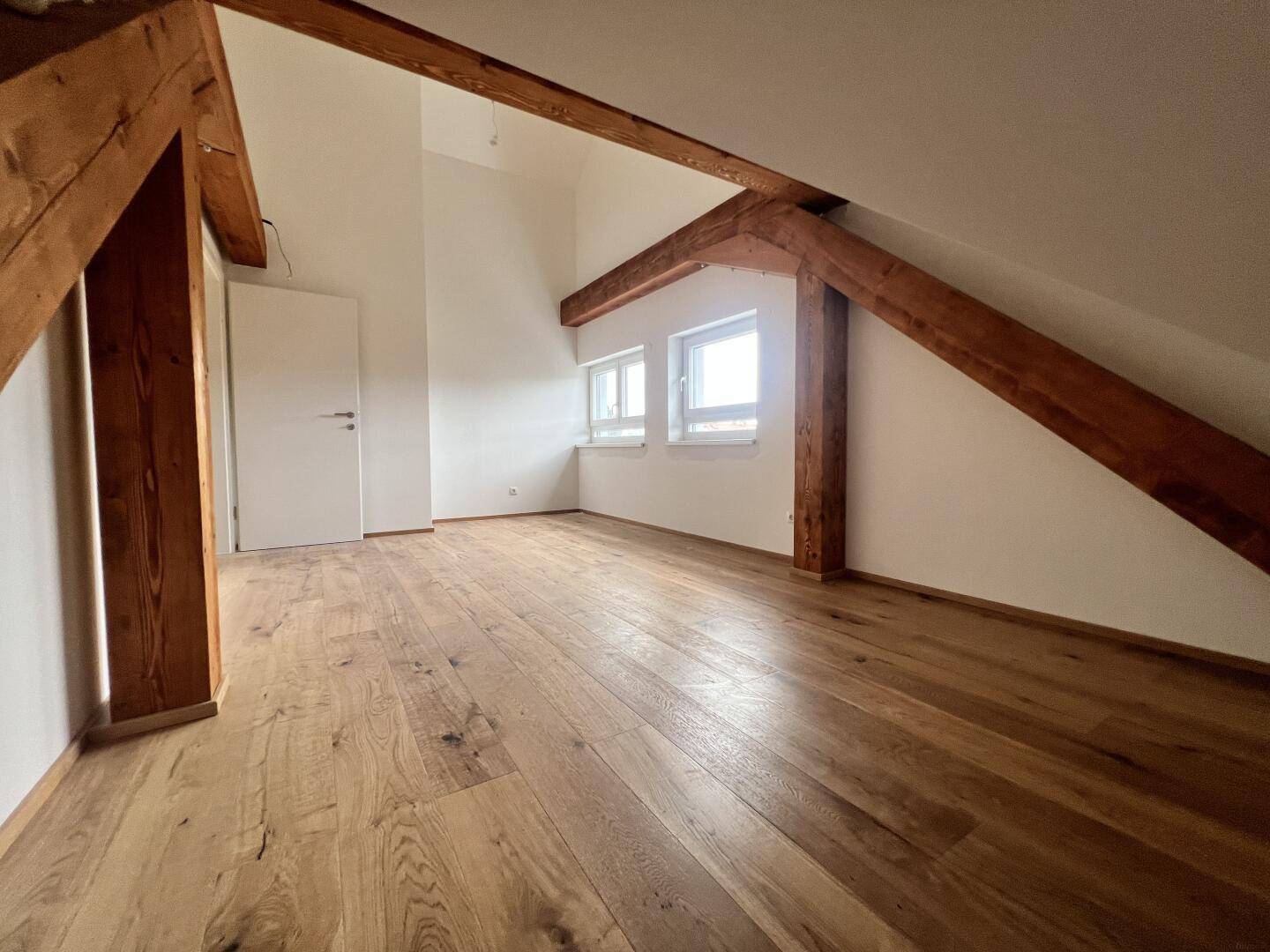 Helles Zimmer mit Holzboden, sichtbaren Balken, Dachschräge und Fenstern.