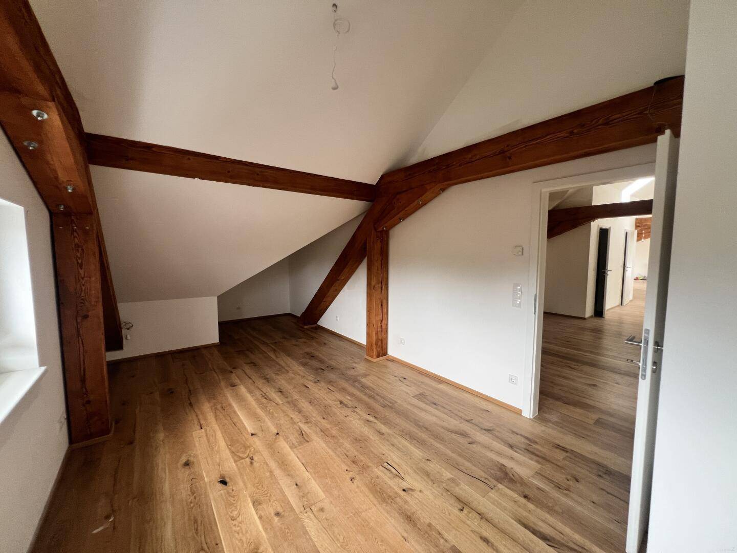 Zimmer mit Holzboden, Holzbalken, Dachschräge und Zugang zu weiteren Räumen.