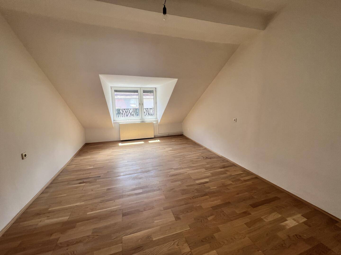 Helles Dachzimmer mit Parkettboden, schrägen Wänden und Fenster.