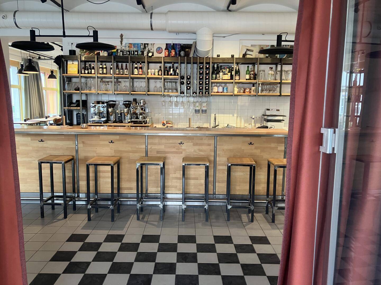 Bar mit Holztresen und Hockern, dahinter Regale mit Flaschen und Gläsern, auf kariertem Fliesenboden.