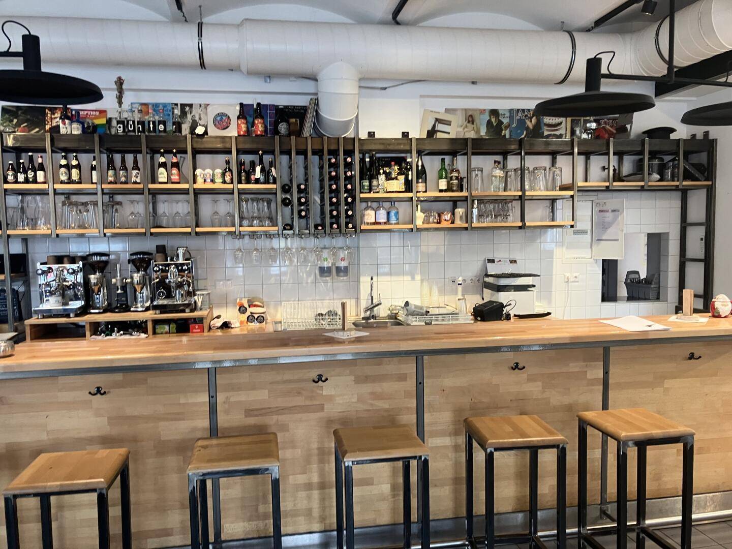 Nahaufnahme der Bar mit Holztresen, Kaffeemaschinen und Regalen voller Getränke und Gläser, gut ausgestattet.