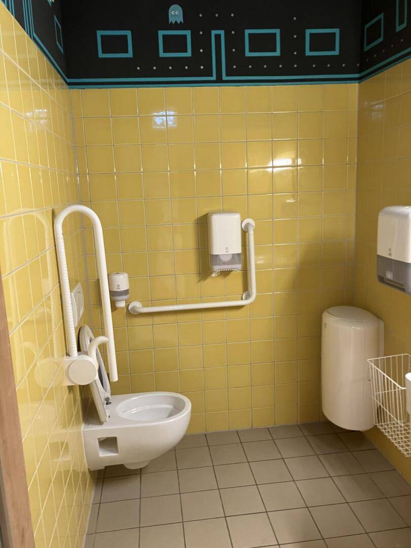 Barrierefreie Toilette mit gelben Fliesen, Haltegriffen und einem wandhängenden WC, Pac-Man-Dekor, modern.