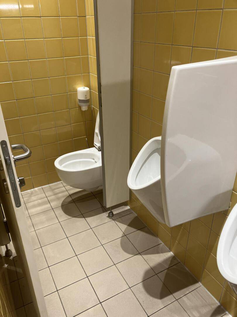 Herrentoilette mit gelben Fliesen, wandhängendem WC und Urinalen, sauber und funktional gestaltet.