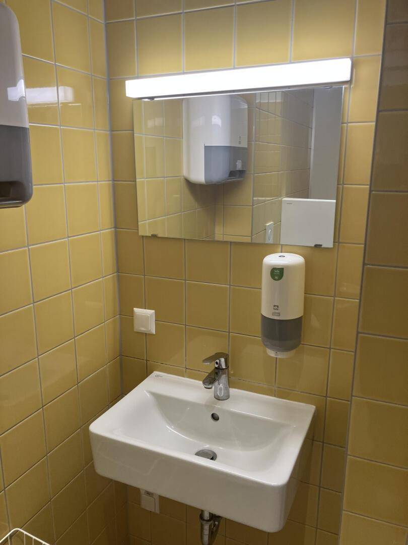 Toilette mit gelben Fliesen, Waschbecken, Spiegel und Seifenspender, helle Beleuchtung, modern und sauber.