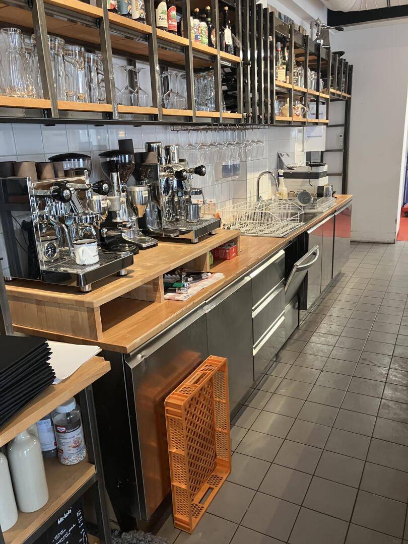 Detailansicht der Bar mit Kaffeemaschinen, Spülbecken und Edelstahloberflächen, dahinter Regale, gut ausgestattet.