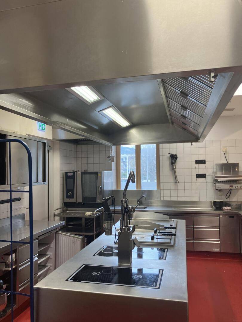 Professionelle Küche mit großem Dunstabzug, Edelstahlgeräten und einem Fenster mit Tageslicht, gut belüftet.