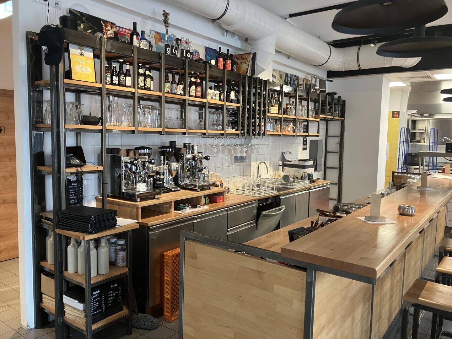 Barbereich mit Holztresen, Kaffeemaschinen und Regalen voller Getränke und Gläser, industrieller Stil, gut ausgestattet.