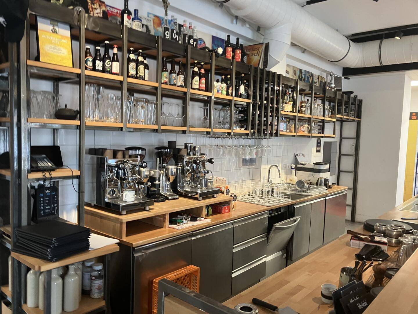 Barbereich mit Holztresen, Kaffeemaschinen und Regalen voller Getränke und Gläser, industrieller Stil, gut ausgestattet.