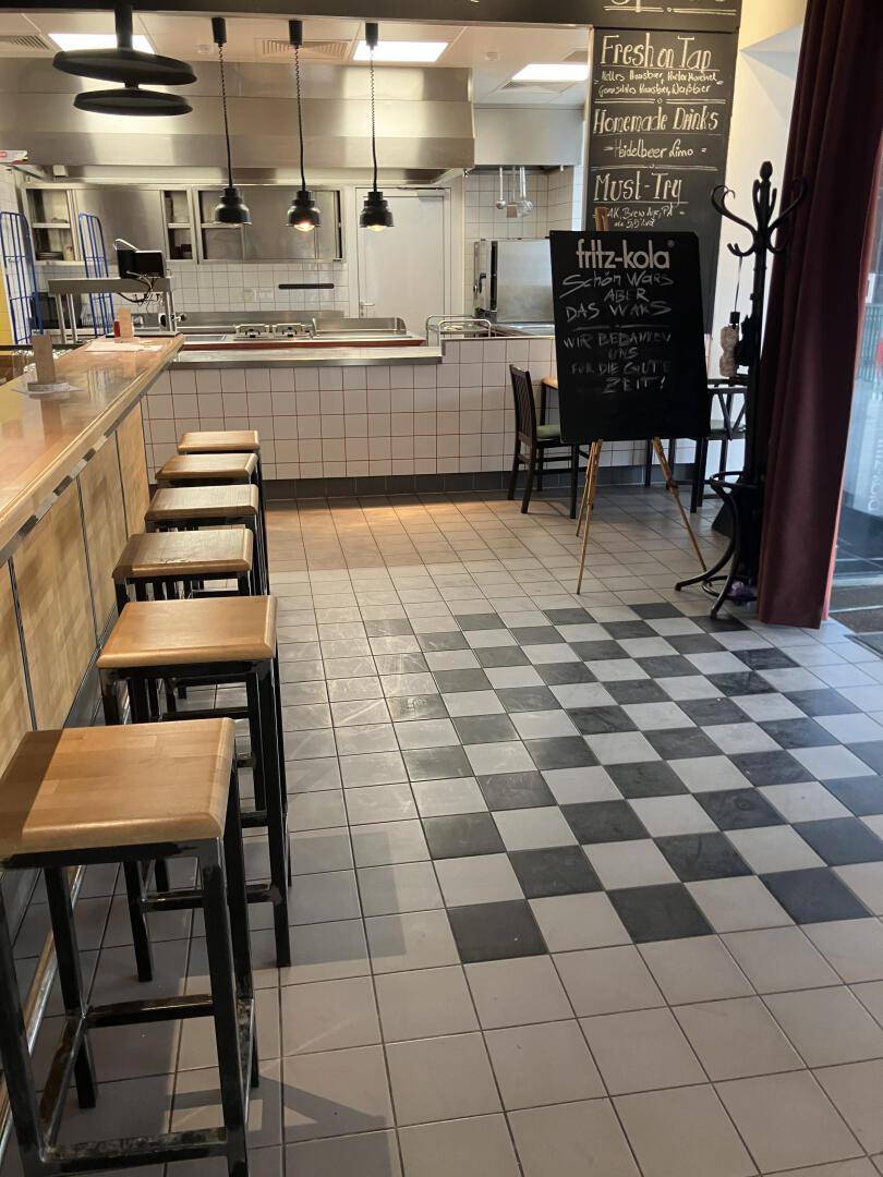 Blick von der Bar in die professionelle Küche mit Edelstahloberflächen und Hängelampen, Klimaanlage sichtbar, modern.