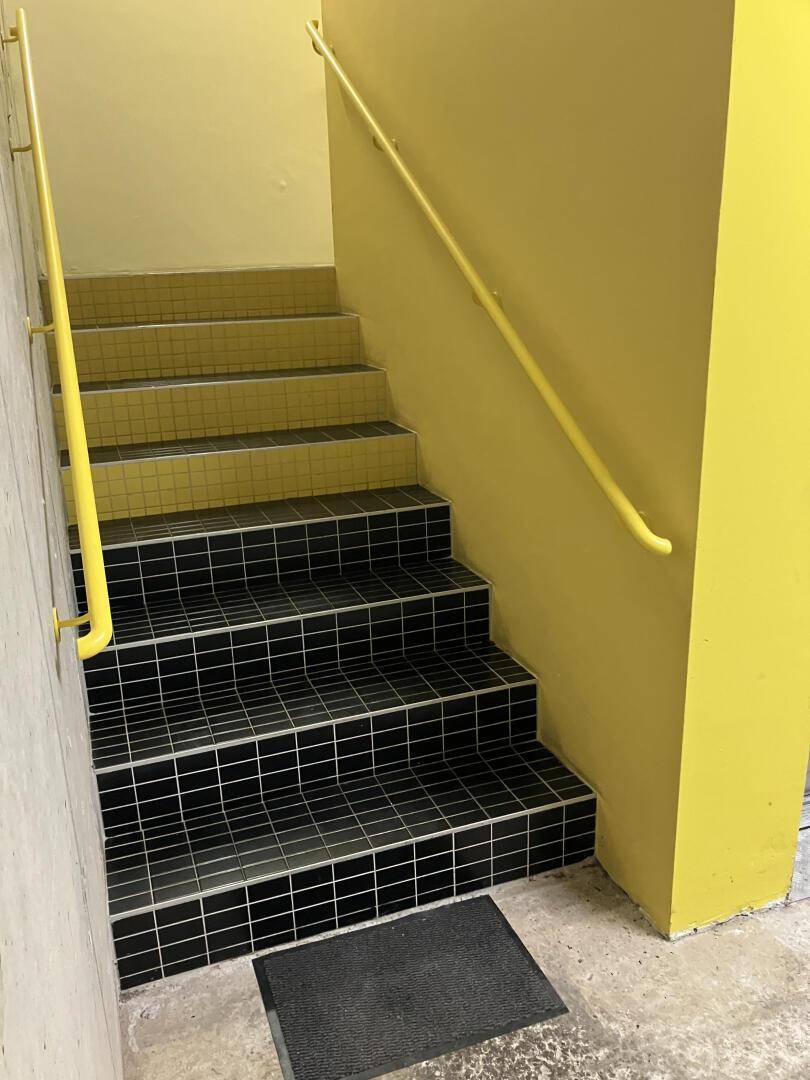 Treppenhaus mit gelben Wänden, schwarzem Fliesenboden und gelbem Handlauf, gut beleuchtet und sicher.