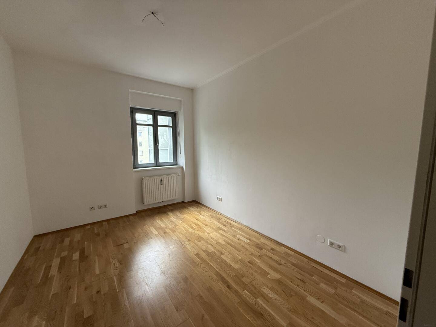 Leeres Zimmer mit Holzboden und einem Fenster, das viel Tageslicht hereinlässt.