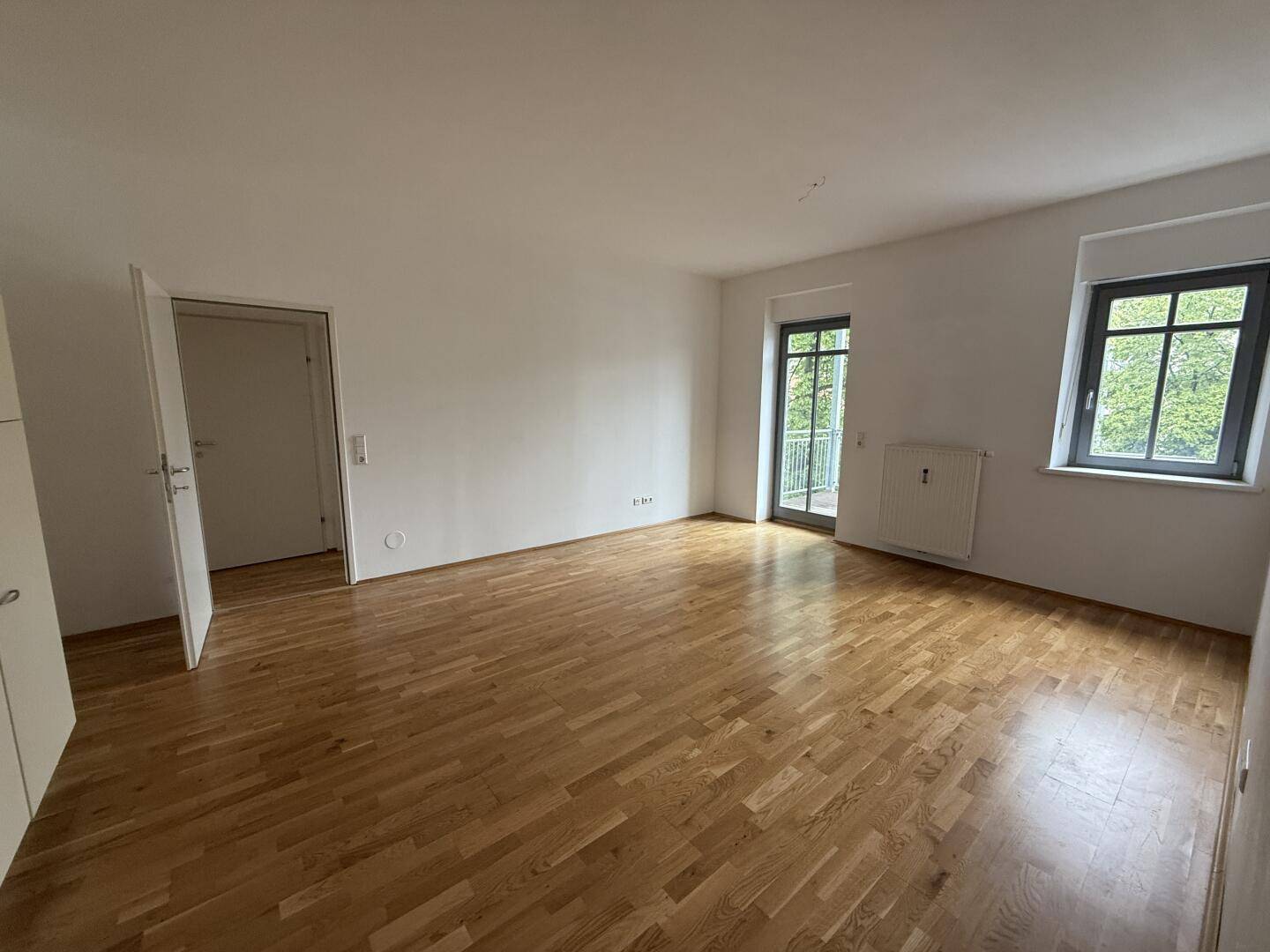 Heller Wohnraum mit schönem Parkettboden, Zugang zum Balkon und einem angenehmen Blick ins Grüne.